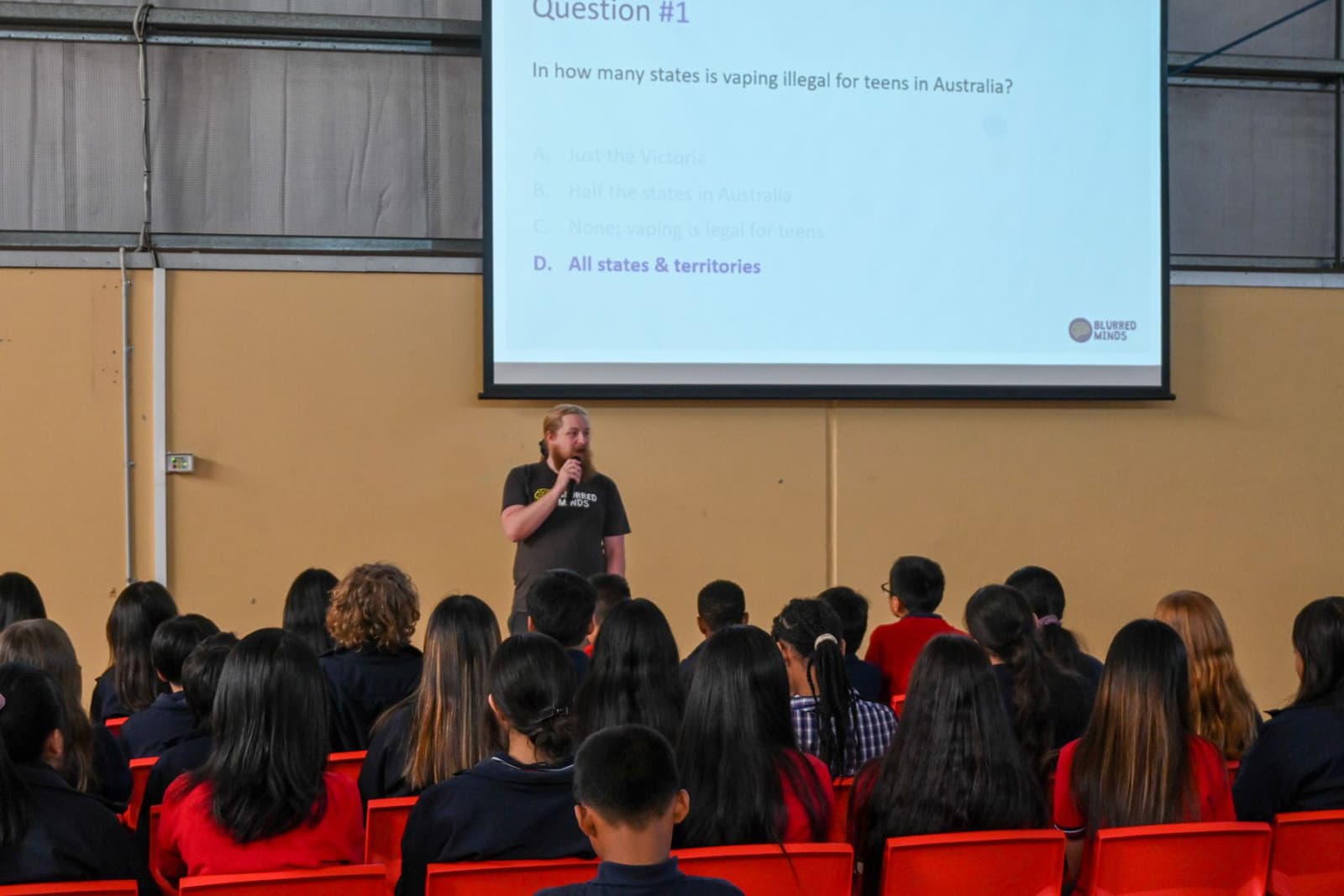 A speaker standing in front of a class of seated students, doing a presentation on drug education.